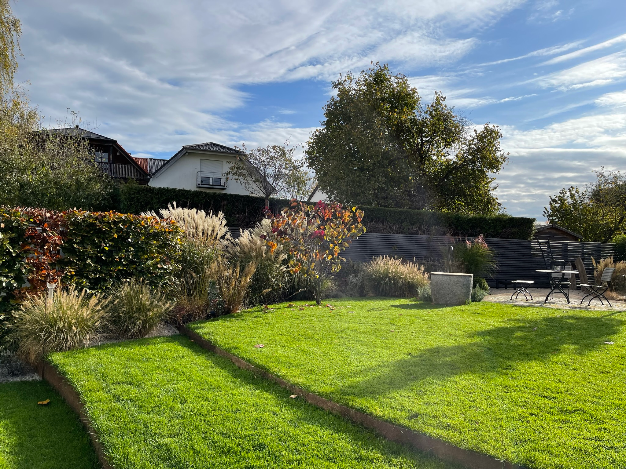 Weite Gartenansicht mit Sitzplatz, Gräserpflanzungen und geschwungenen Cortenstahlbändern